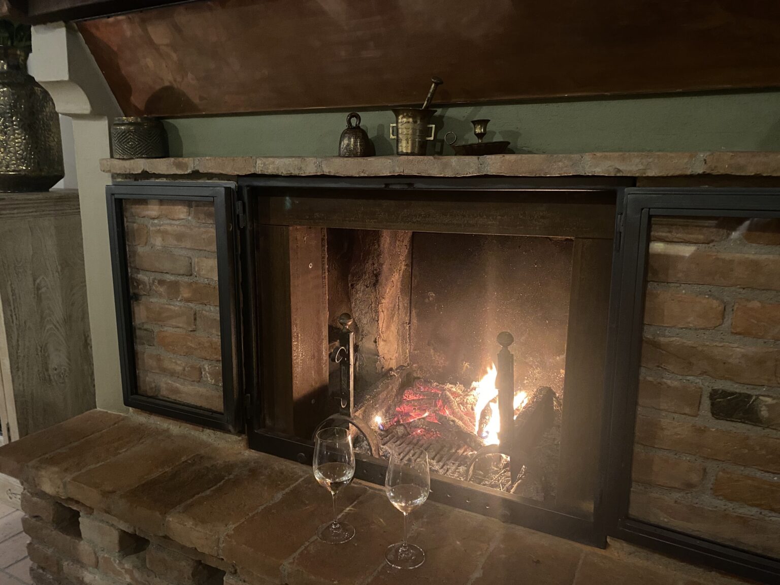 Cozy fireplace with a glowing fire, surrounded by rustic brick and a wooden mantel, featuring two wine glasses on the stone hearth, creating a warm and inviting atmosphere. | Only on SopranoVillas