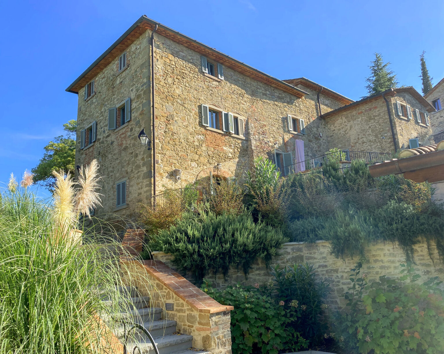 Stone house with green shutters surrounded by lush gardens and stairs leading to the entrance, under a clear blue sky. | Only on SopranoVillas