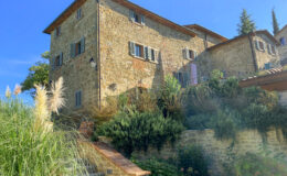 Stone house with green shutters surrounded by lush gardens and stairs leading to the entrance, under a clear blue sky.