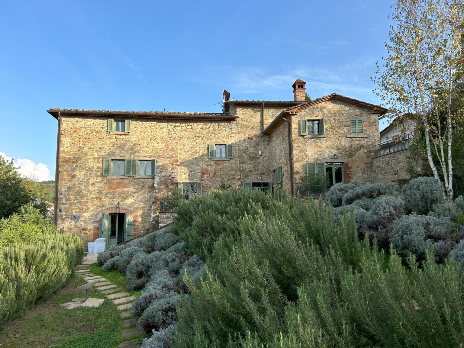 Rustic stone villa surrounded by lush greenery and lavender plants, featuring green shutters and a blue sky in the background. Ideal for vacation rentals in a serene countryside setting. | Only on SopranoVillas