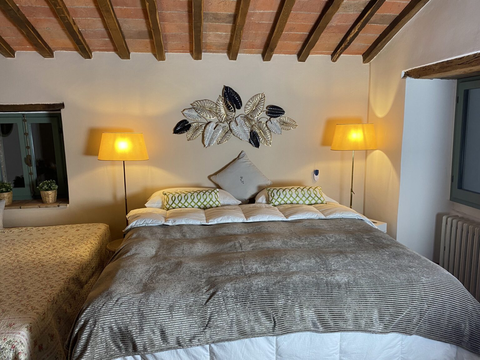 Cozy bedroom interior featuring a large bed with a soft gray blanket, decorative pillows, and stylish leaf wall art. Two warm lamps illuminate the space, complemented by a window with green accents and potted plants. The room exudes a warm and inviting atmosphere, perfect for relaxation. | Only on SopranoVillas