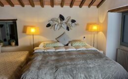 Cozy bedroom interior featuring a large bed with a soft gray blanket, decorative pillows, and stylish leaf wall art. Two warm lamps illuminate the space, complemented by a window with green accents and potted plants. The room exudes a warm and inviting atmosphere, perfect for relaxation.