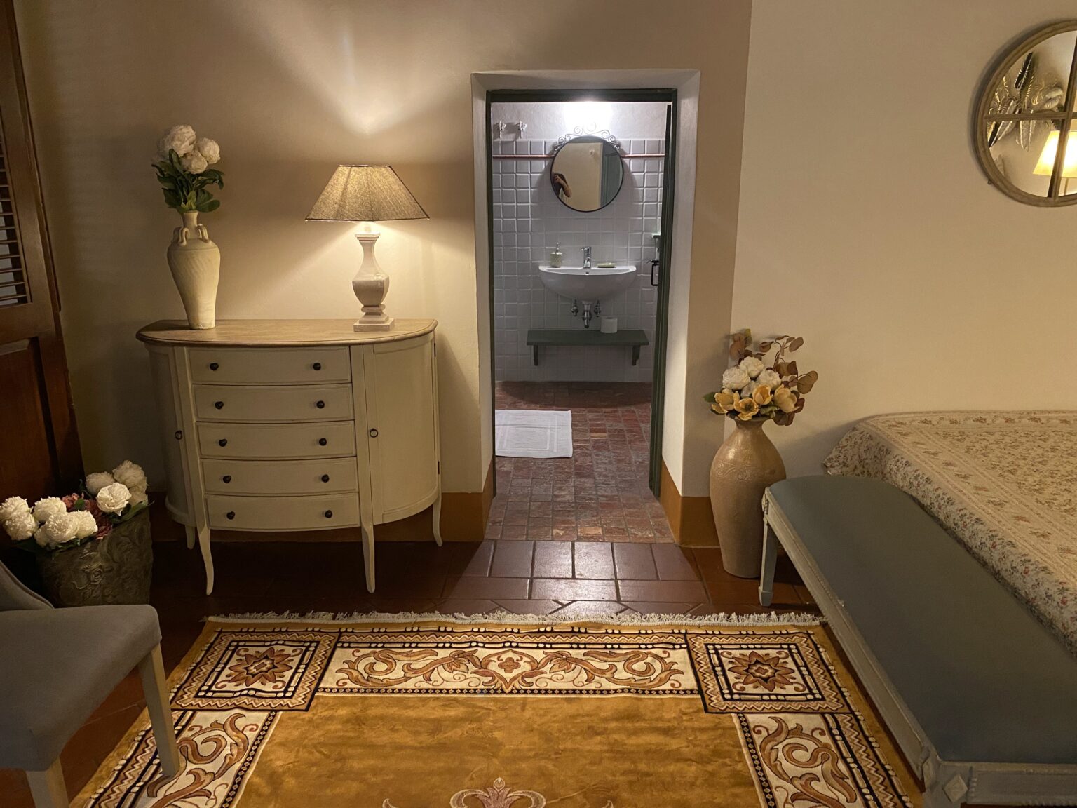 Cozy interior view of a room featuring a stylish dresser with decorative flowers, a lamp, and a comfortable seating area. The room leads to a bathroom with a sink and mirror, showcasing warm lighting and elegant decor elements like a patterned rug and a vase of flowers. | Only on SopranoVillas