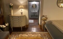 Cozy interior view of a room featuring a stylish dresser with decorative flowers, a lamp, and a comfortable seating area. The room leads to a bathroom with a sink and mirror, showcasing warm lighting and elegant decor elements like a patterned rug and a vase of flowers.