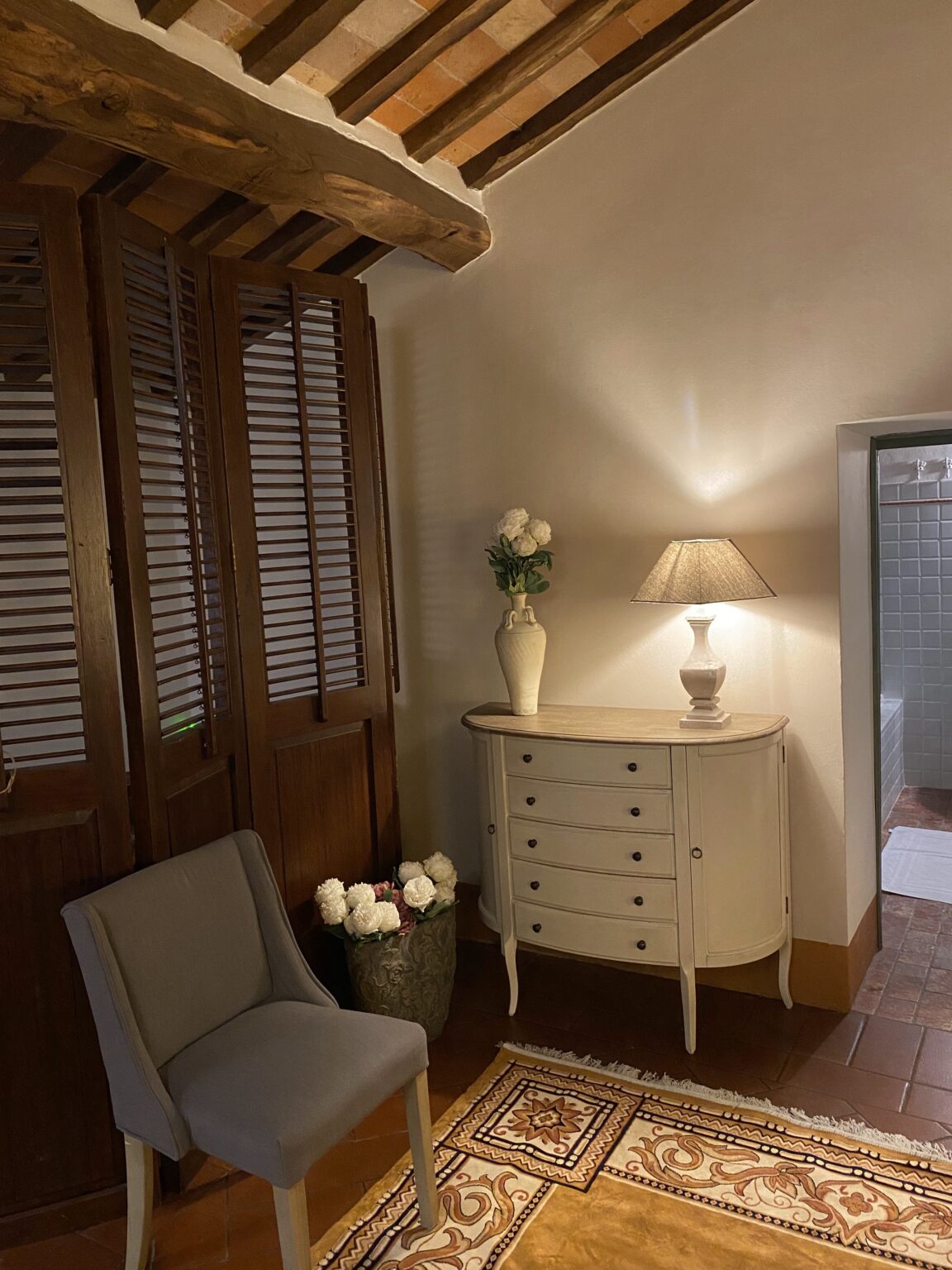 Cozy interior design featuring a gray chair, a vintage-style dresser, and decorative flowers in a vase. The room has a warm ambiance with wooden beams and a soft lamp light, enhancing the inviting atmosphere. A patterned rug adds elegance to the space, while a glimpse of a bathroom is visible in the background. | Only on SopranoVillas