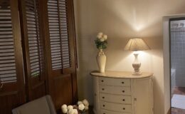Cozy interior design featuring a gray chair, a vintage-style dresser, and decorative flowers in a vase. The room has a warm ambiance with wooden beams and a soft lamp light, enhancing the inviting atmosphere. A patterned rug adds elegance to the space, while a glimpse of a bathroom is visible in the background.