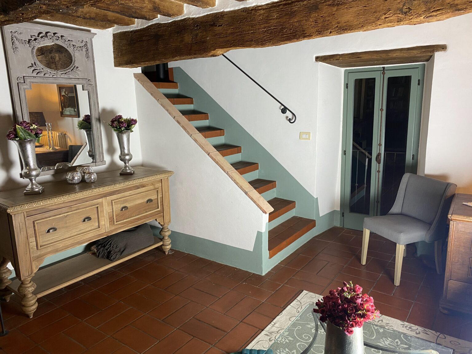 Cozy interior of a rustic home featuring a wooden staircase, vintage mirror, and decorative vases with flowers on a console table. A gray chair and a glass door are visible, enhancing the inviting atmosphere. | Only on SopranoVillas