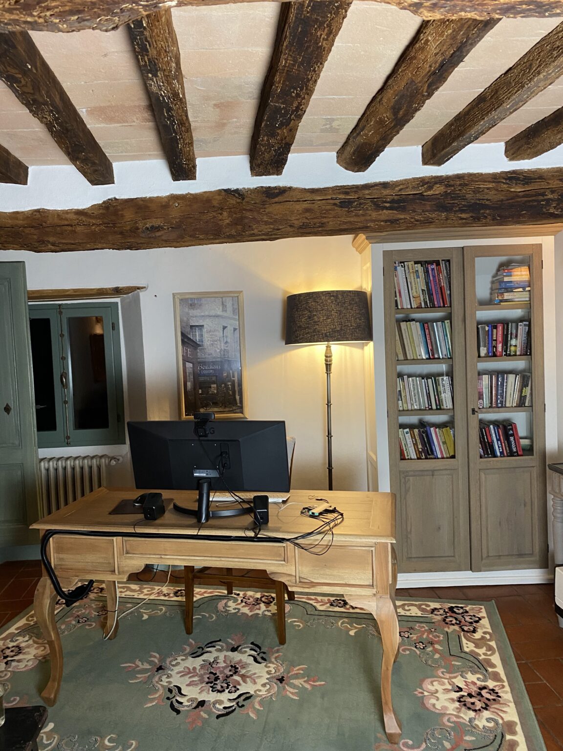 Home office setup featuring a wooden desk with a computer monitor, lamp, and various office supplies. The space includes a bookshelf filled with books, rustic wooden beams overhead, and a decorative rug, creating a cozy and inviting atmosphere. | Only on SopranoVillas
