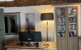 Home office setup featuring a wooden desk with a computer monitor, lamp, and various office supplies. The space includes a bookshelf filled with books, rustic wooden beams overhead, and a decorative rug, creating a cozy and inviting atmosphere.