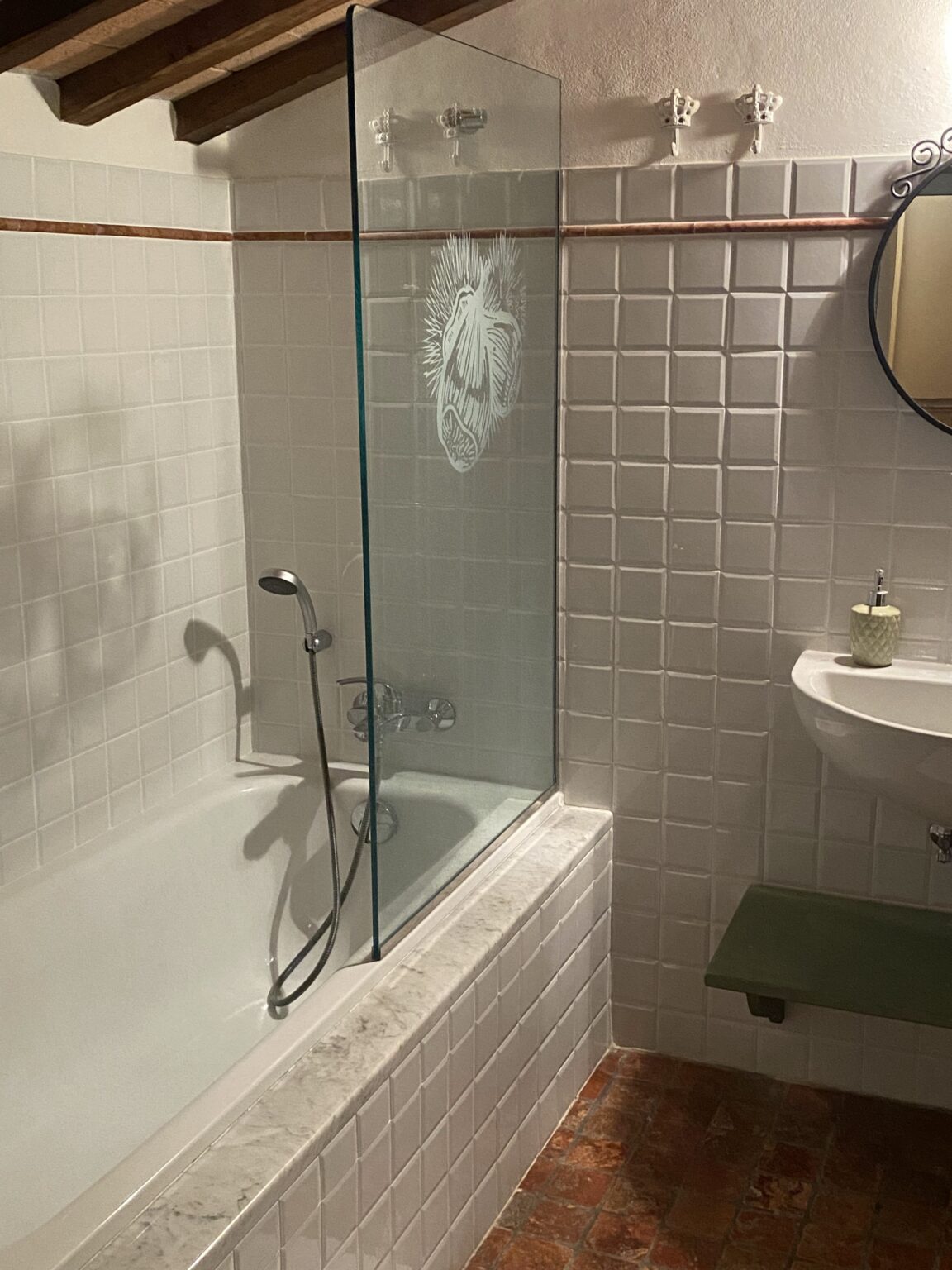 Modern bathroom featuring a glass shower enclosure with an artistic design, a white tiled bathtub, and a sleek sink. The room has rustic wooden beams and terracotta flooring, creating a warm and inviting atmosphere. | Only on SopranoVillas