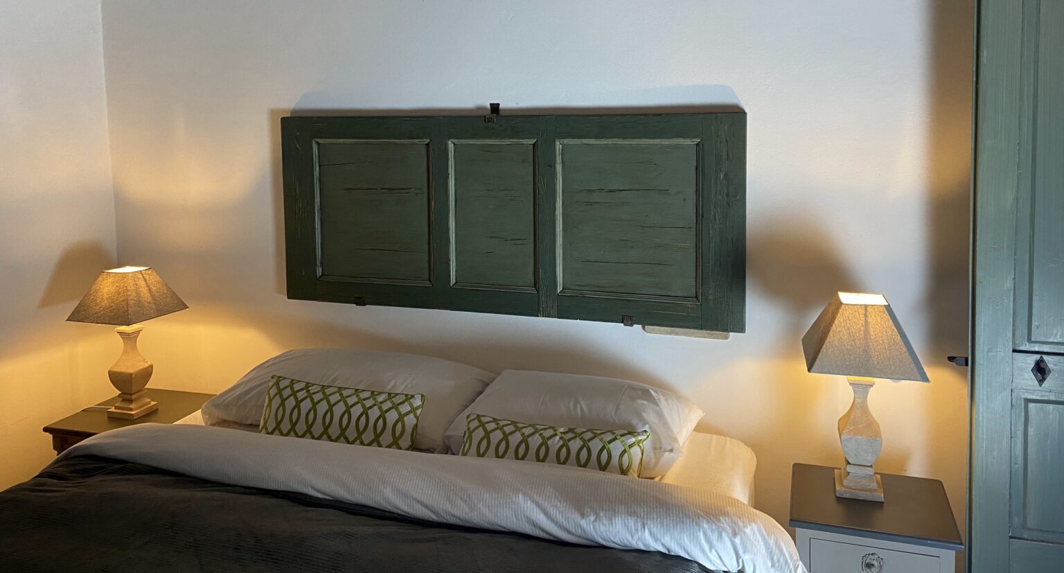 Cozy bedroom featuring a green wooden headboard, two bedside lamps with warm lighting, and decorative pillows on a neatly made bed. The walls are painted white, enhancing the room's bright and inviting atmosphere. | Only on SopranoVillas