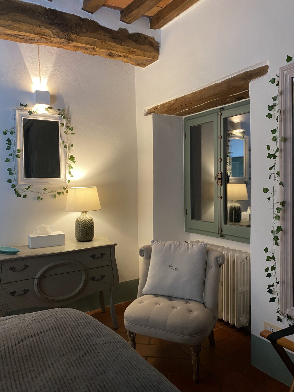 Cozy bedroom interior featuring a decorative mirror with ivy, a soft armchair with a cushion, a stylish lamp, and a vintage dresser, accentuated by wooden beams and warm lighting. | Only on SopranoVillas