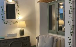 Cozy bedroom interior featuring a decorative mirror with ivy, a soft armchair with a cushion, a stylish lamp, and a vintage dresser, accentuated by wooden beams and warm lighting.