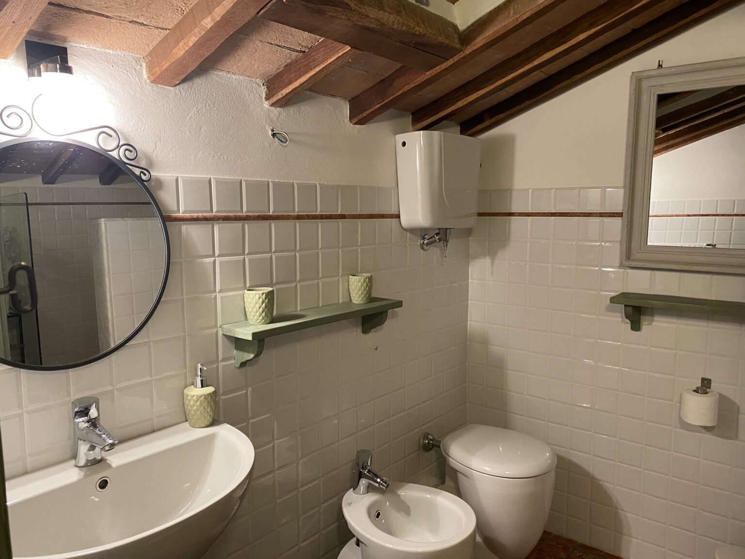 Modern bathroom interior featuring a round mirror, sink, toilet, and bidet, with white tiled walls and wooden beams. Green shelves hold decorative items, adding a touch of style to the space. | Only on SopranoVillas