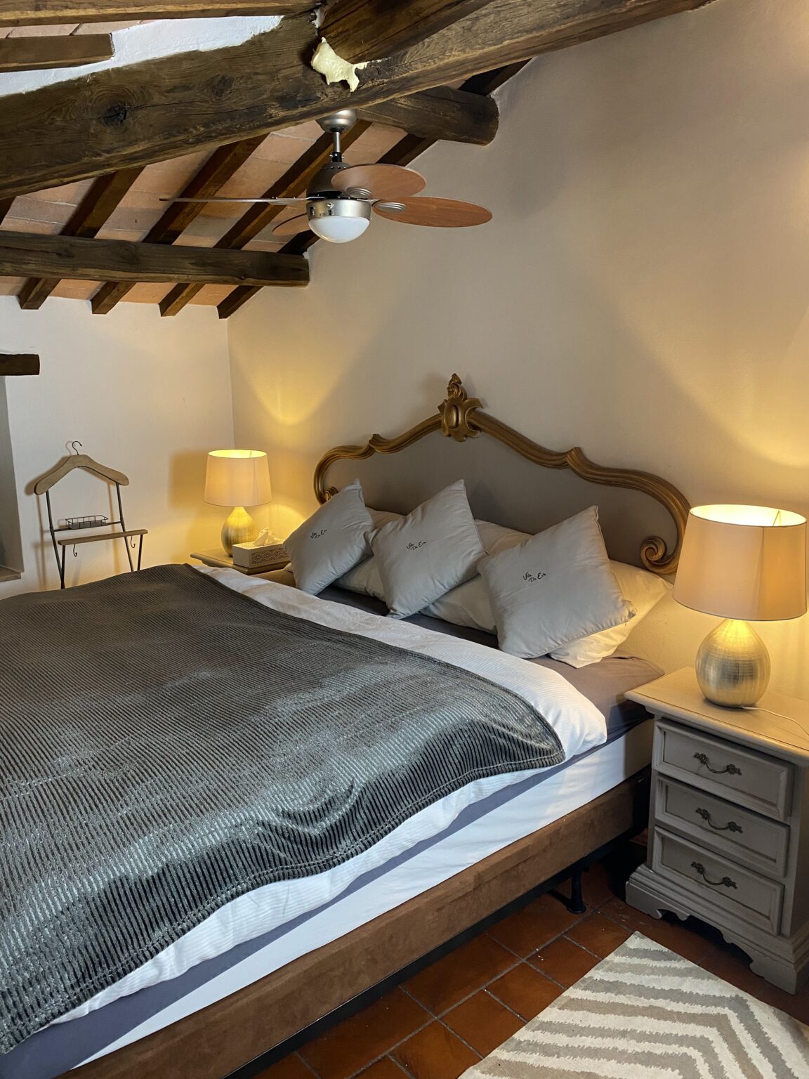 Cozy bedroom interior featuring a wooden ceiling with exposed beams, a stylish bed with a gray blanket and multiple pillows, two bedside lamps providing warm lighting, and a decorative rug on the floor. Ideal for a relaxing retreat. | Only on SopranoVillas