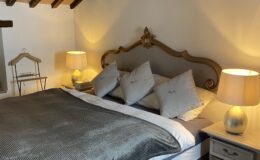 Cozy bedroom interior featuring a wooden ceiling with exposed beams, a stylish bed with a gray blanket and multiple pillows, two bedside lamps providing warm lighting, and a decorative rug on the floor. Ideal for a relaxing retreat.