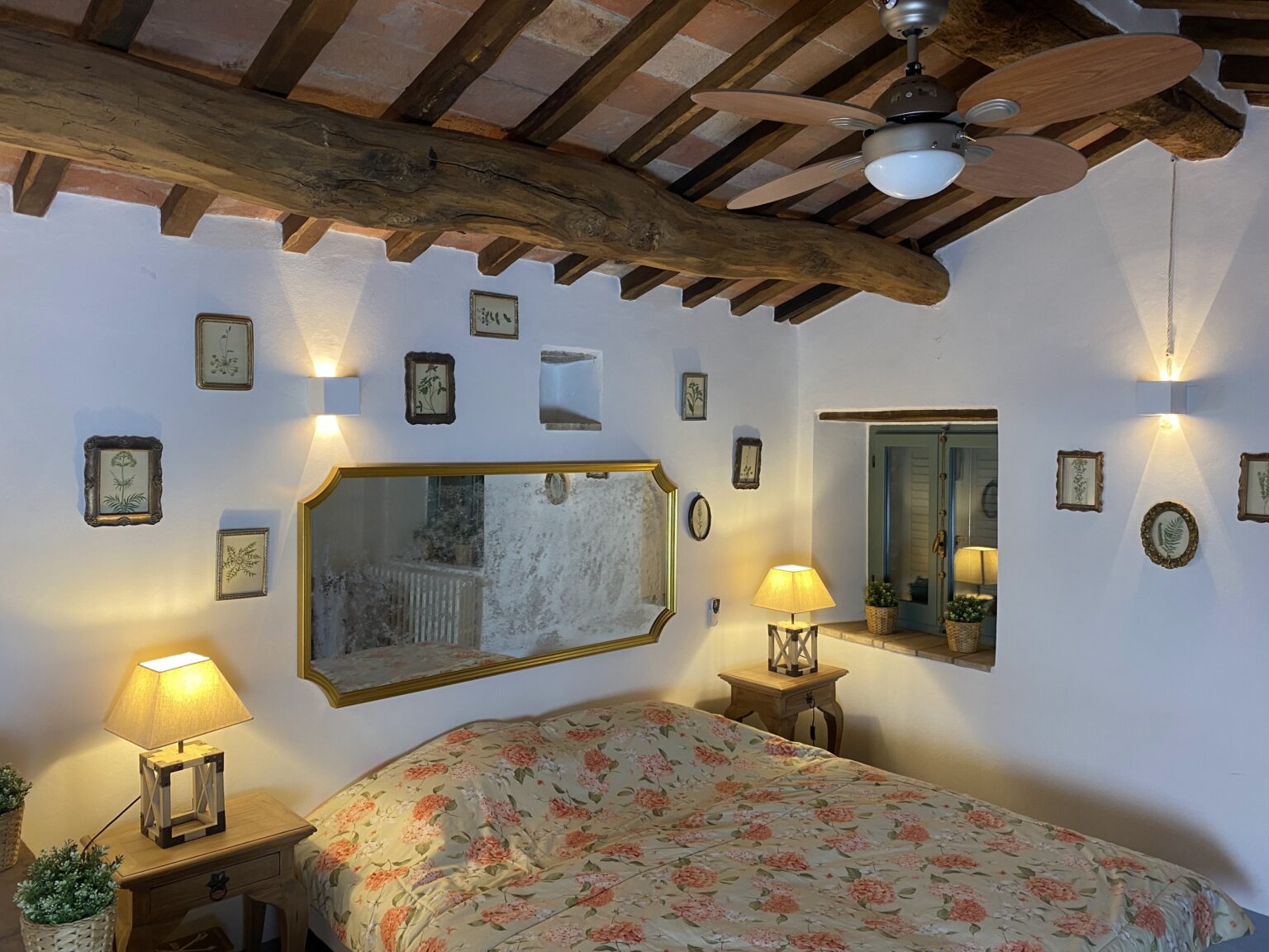 Cozy bedroom featuring a floral bedspread, wooden beams, and decorative wall art. The room includes a mirror, bedside lamps, and a window with potted plants, creating a warm and inviting atmosphere. Perfect for relaxation and comfort. | Only on SopranoVillas