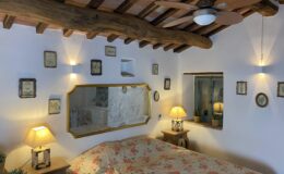 Cozy bedroom featuring a floral bedspread, wooden beams, and decorative wall art. The room includes a mirror, bedside lamps, and a window with potted plants, creating a warm and inviting atmosphere. Perfect for relaxation and comfort.
