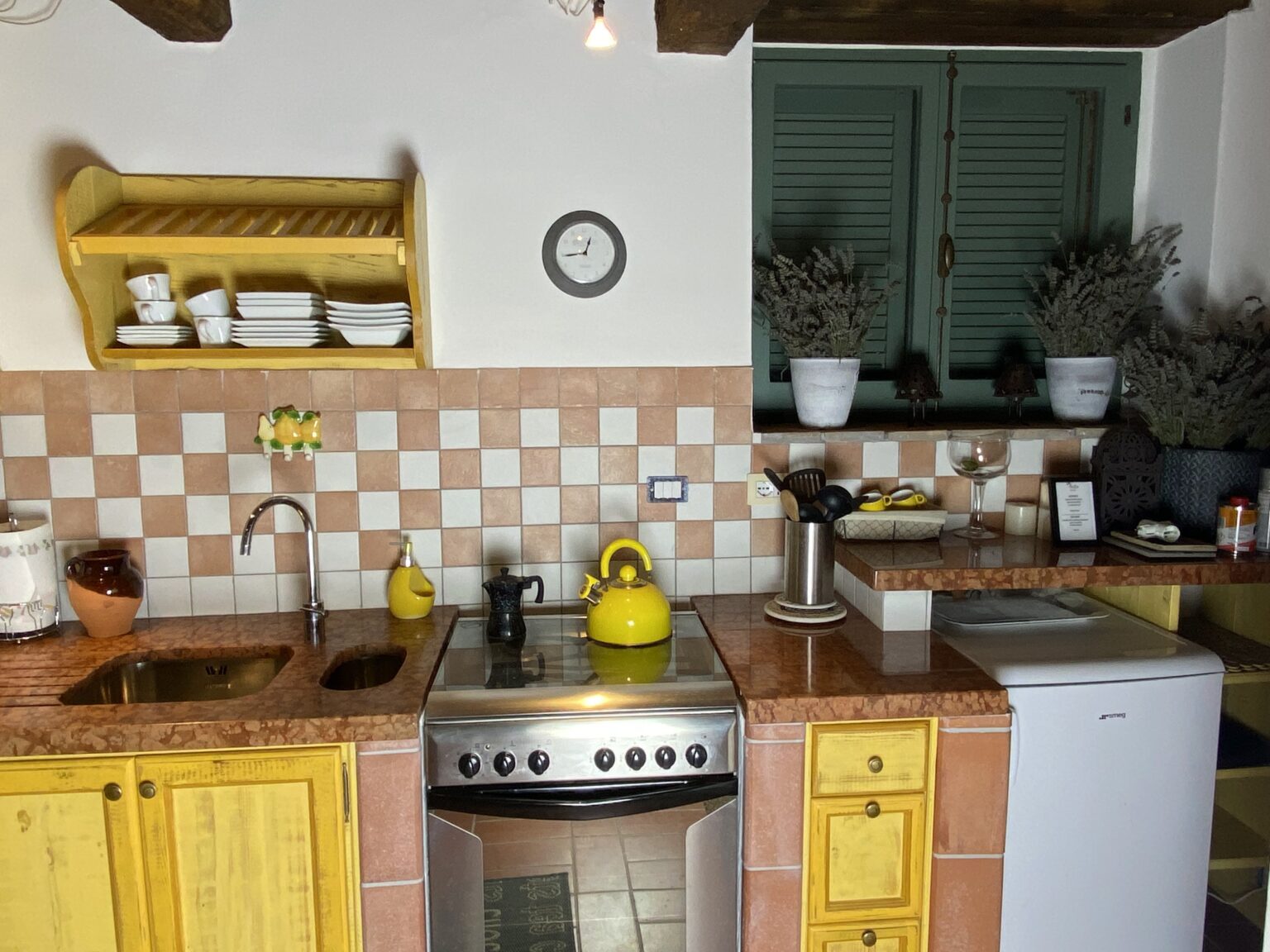 Cozy kitchen interior featuring yellow cabinets, a stainless steel stove, and a double sink. The backsplash is a checkerboard pattern of peach and white tiles. A wooden shelf displays white dishes, and potted plants add a decorative touch to the space. A vintage clock and yellow kettle enhance the homey atmosphere. | Only on SopranoVillas