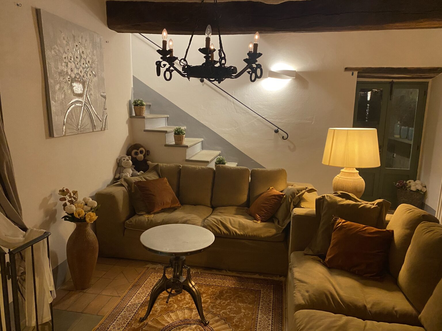 Cozy living room with a beige sofa adorned with decorative pillows, a round coffee table, and a warm floor lamp. The wall features a bicycle-themed artwork, and a staircase leads to an upper level, decorated with potted plants and plush toys. The space is illuminated by a stylish chandelier, creating a welcoming atmosphere. | Only on SopranoVillas