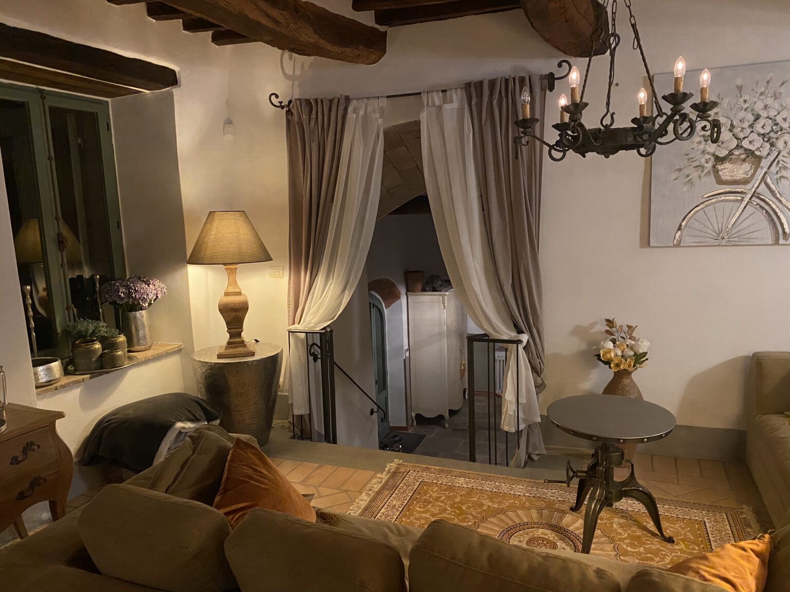 Cozy living area featuring rustic decor, a warm lamp, elegant curtains, and a stylish chandelier, with a staircase leading to an upper level. The space is adorned with a decorative vase and comfortable seating, creating an inviting atmosphere. | Only on SopranoVillas