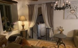 Cozy living area featuring rustic decor, a warm lamp, elegant curtains, and a stylish chandelier, with a staircase leading to an upper level. The space is adorned with a decorative vase and comfortable seating, creating an inviting atmosphere.