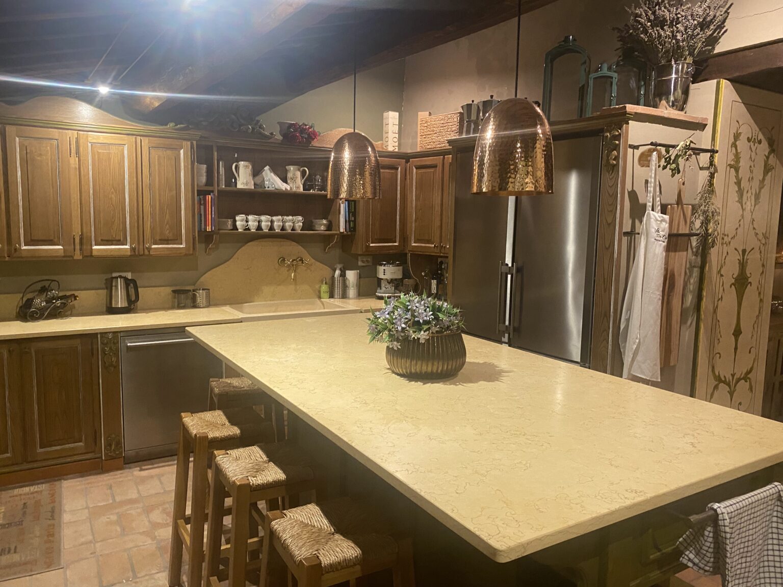 Cozy kitchen interior featuring wooden cabinets, a large countertop, and copper pendant lights. The space includes a stainless steel refrigerator, a kitchen sink, and decorative items like potted plants and dishware. Natural stone flooring adds warmth to the design. | Only on SopranoVillas