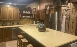 Cozy kitchen interior featuring wooden cabinets, a large countertop, and copper pendant lights. The space includes a stainless steel refrigerator, a kitchen sink, and decorative items like potted plants and dishware. Natural stone flooring adds warmth to the design.