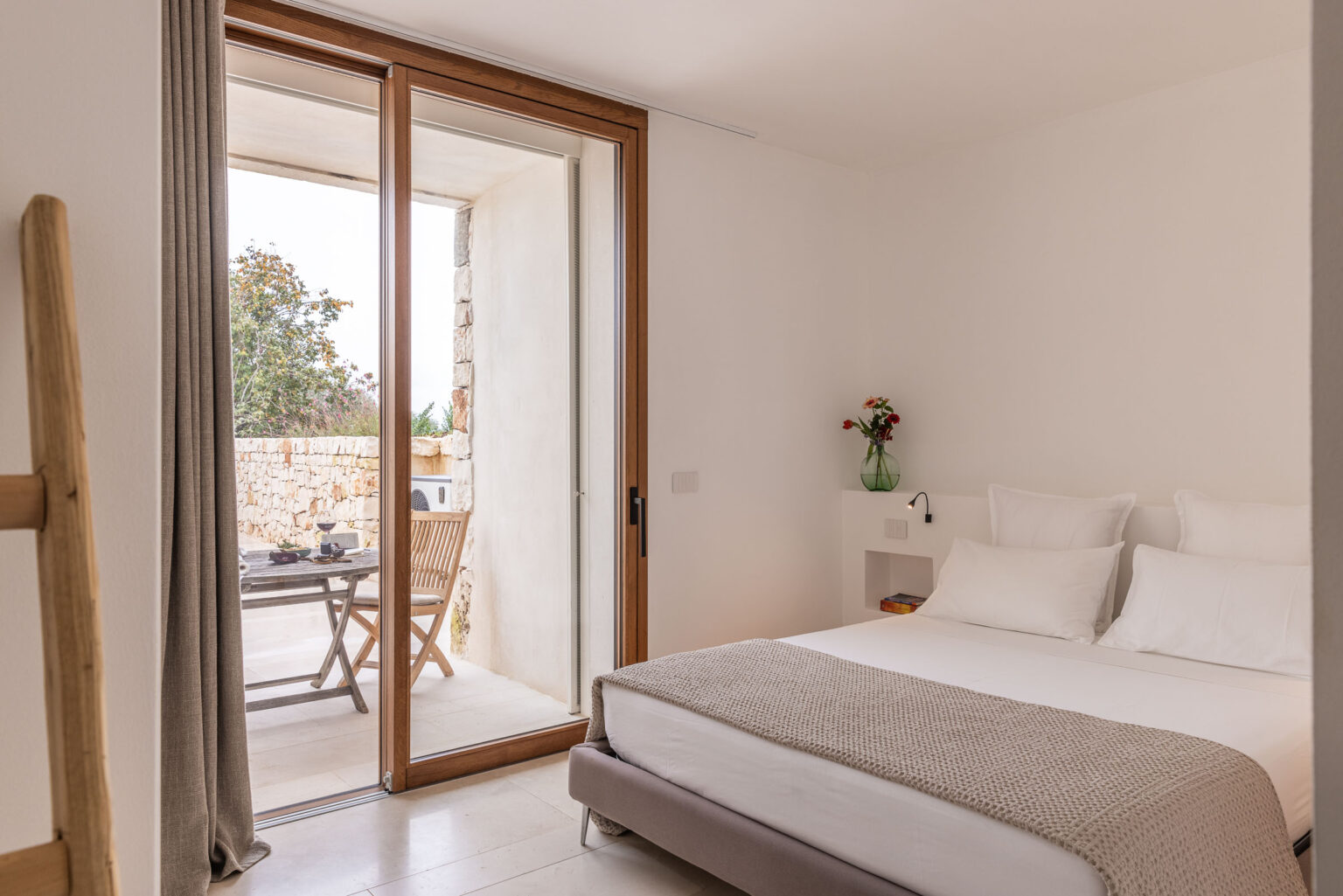Modern bedroom featuring a cozy bed with white linens and a textured throw blanket, large glass doors leading to a stone patio with outdoor seating, and minimalist decor with natural light. | Only on SopranoVillas