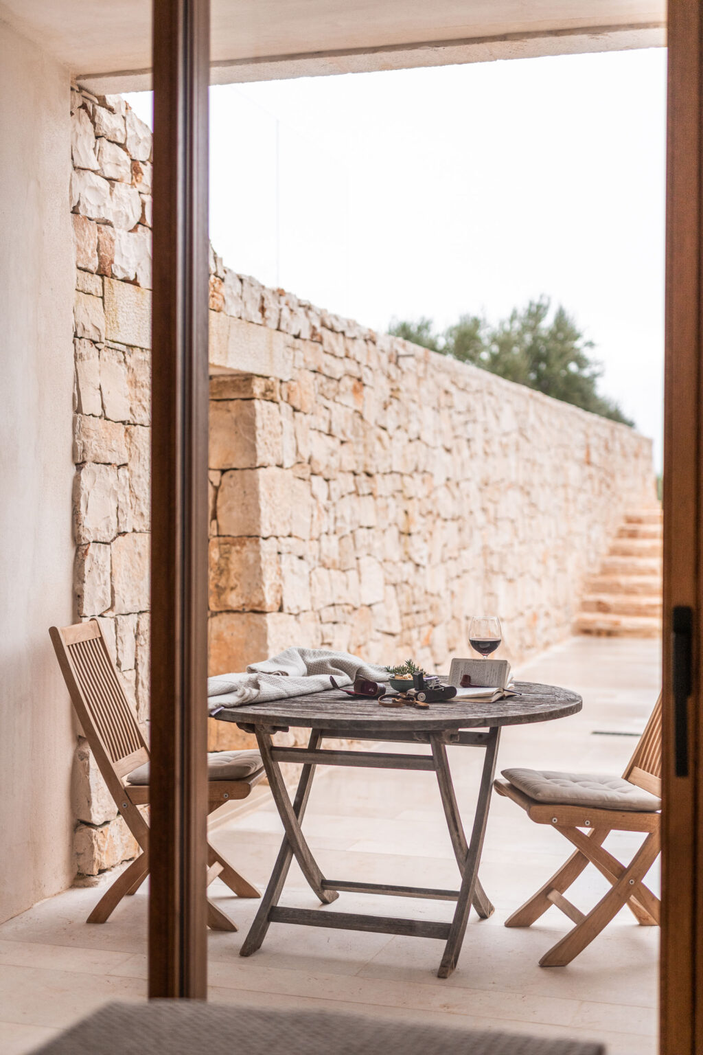 Cozy outdoor dining area featuring a rustic wooden table set with a glass of red wine, a book, and decorative items, framed by a natural stone wall and lush greenery in the background. Ideal for relaxation and enjoying the outdoors. | Only on SopranoVillas