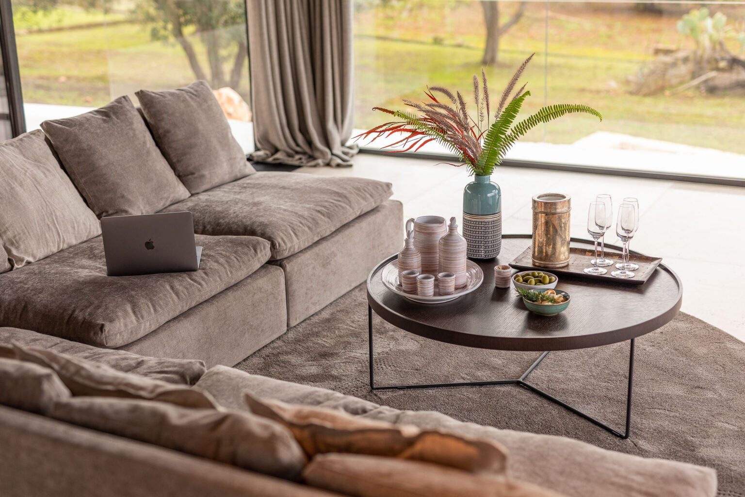Modern living room with a cozy beige sofa, a round coffee table featuring decorative items, snacks, and glasses, complemented by large windows showcasing a scenic outdoor view. | Only on SopranoVillas