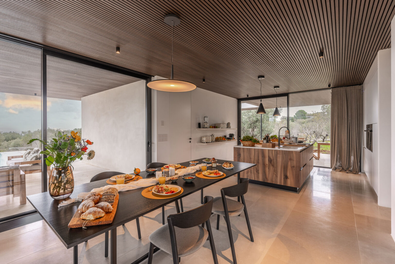 Modern kitchen interior featuring a wooden dining table set with breakfast dishes, fresh flowers in a vase, and a spacious open layout. The kitchen area boasts sleek cabinetry and large windows that provide natural light and views of the outdoor landscape. | Only on SopranoVillas