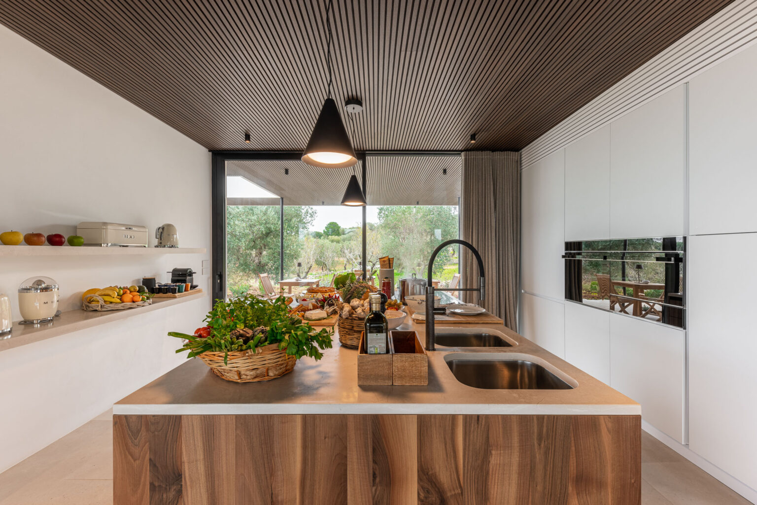 Modern kitchen interior featuring a wooden island with fresh vegetables and fruits, sleek appliances, and large windows overlooking a garden. The design includes wooden ceiling panels and minimalistic cabinetry, creating a bright and inviting atmosphere. | Only on SopranoVillas