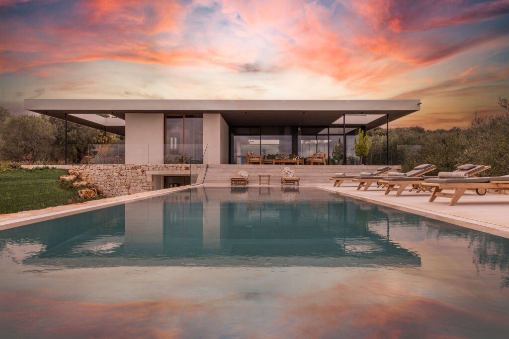 Modern villa with a sleek design, featuring a swimming pool in the foreground and a stunning sunset sky in the background. The property is surrounded by lush greenery and outdoor lounge chairs, creating a serene and inviting atmosphere. | Only on SopranoVillas