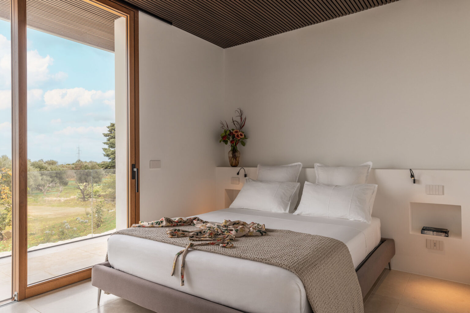 Modern bedroom with a double bed adorned with decorative pillows and a cozy throw, featuring large glass doors that open to a scenic outdoor view. The room has minimalist decor, including a small flower arrangement and built-in bedside lighting for a stylish ambiance. | Only on SopranoVillas