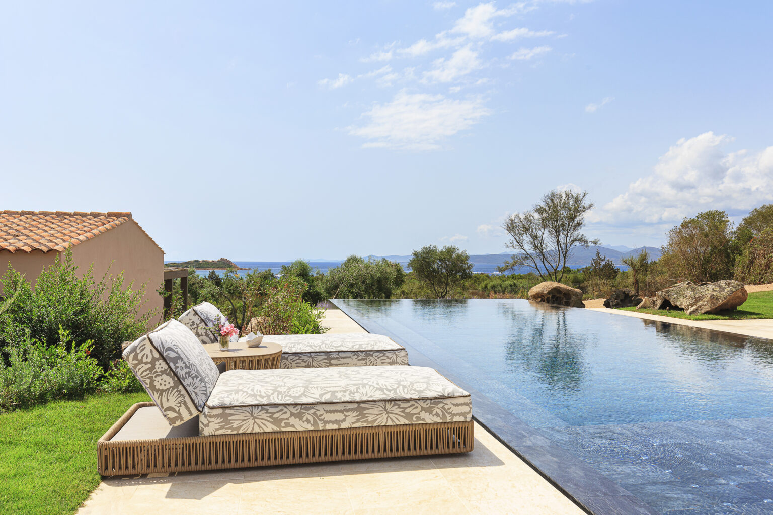 Luxurious outdoor lounge area with two stylish sunbeds beside an infinity pool, overlooking a scenic view of the ocean and lush greenery under a clear blue sky. Ideal for relaxation and enjoying beautiful landscapes. | Only on SopranoVillas