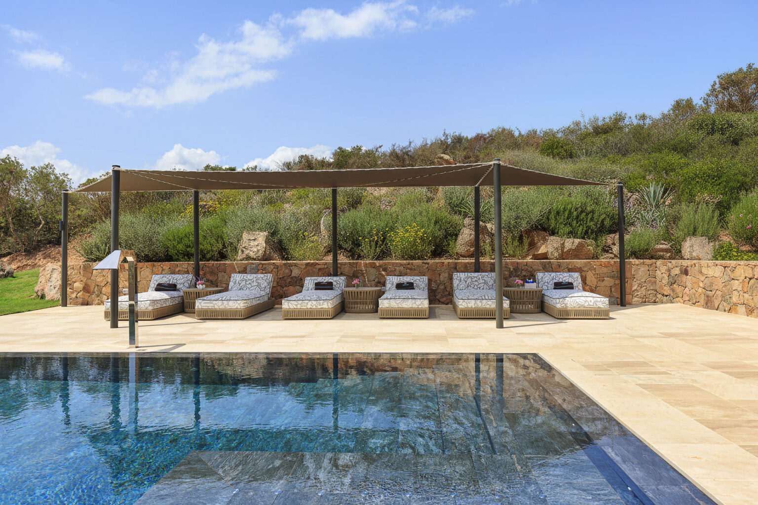 Modern outdoor pool area featuring stylish lounge chairs under a shaded canopy, surrounded by lush greenery and a stone wall, with a clear blue sky reflecting in the water. Ideal for relaxation and leisure. | Only on SopranoVillas