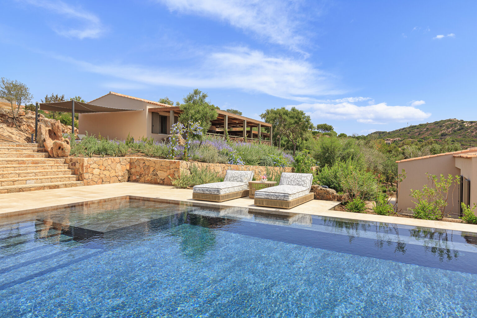 Luxury villa with an infinity pool surrounded by lush greenery and stone pathways, featuring sun loungers and a modern architectural design against a clear blue sky. Ideal for relaxation and outdoor living. | Only on SopranoVillas