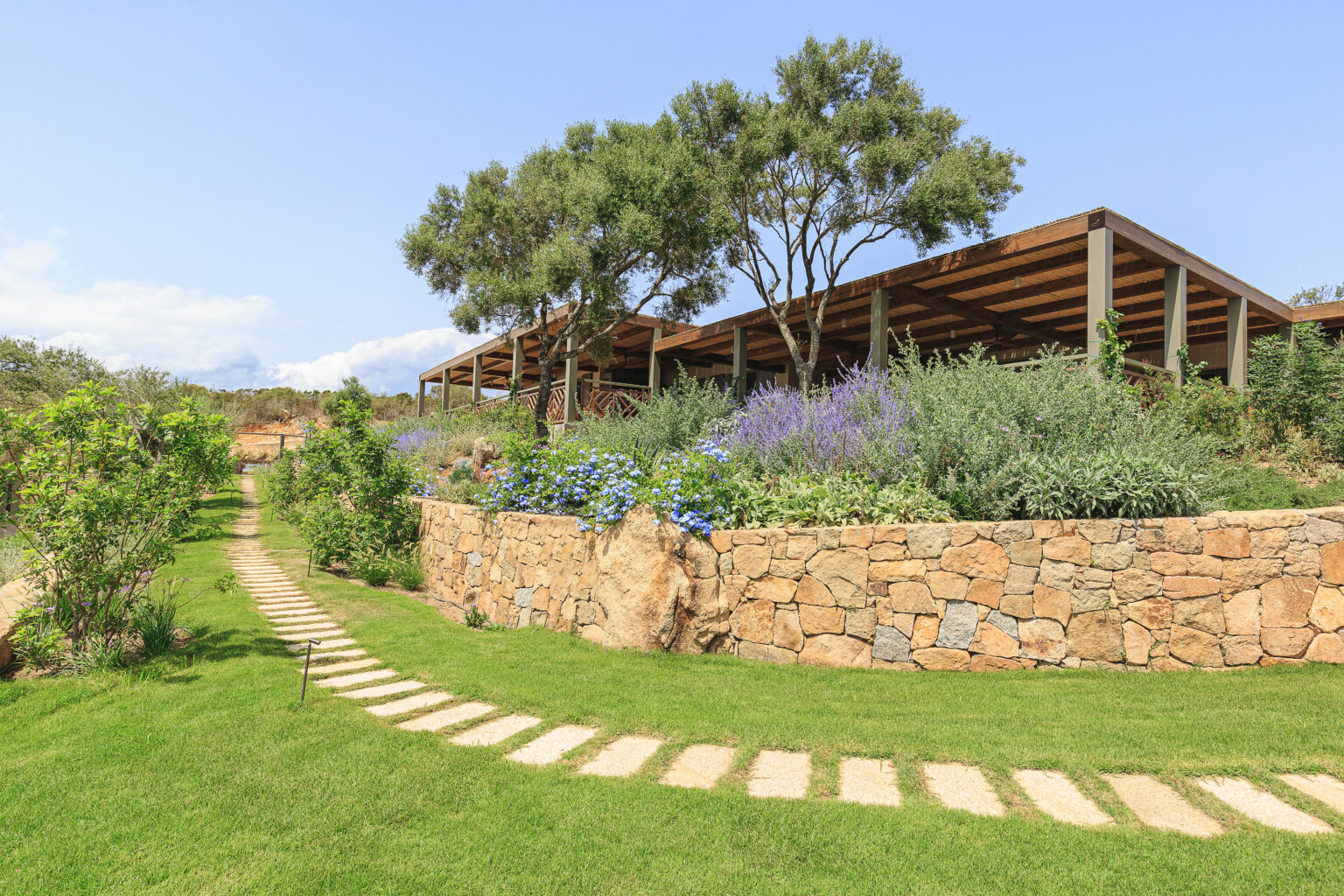 Scenic landscape featuring a stone pathway leading through lush greenery and colorful flowers, with a modern building nestled among trees under a clear blue sky. Ideal for nature lovers and outdoor enthusiasts. | Only on SopranoVillas