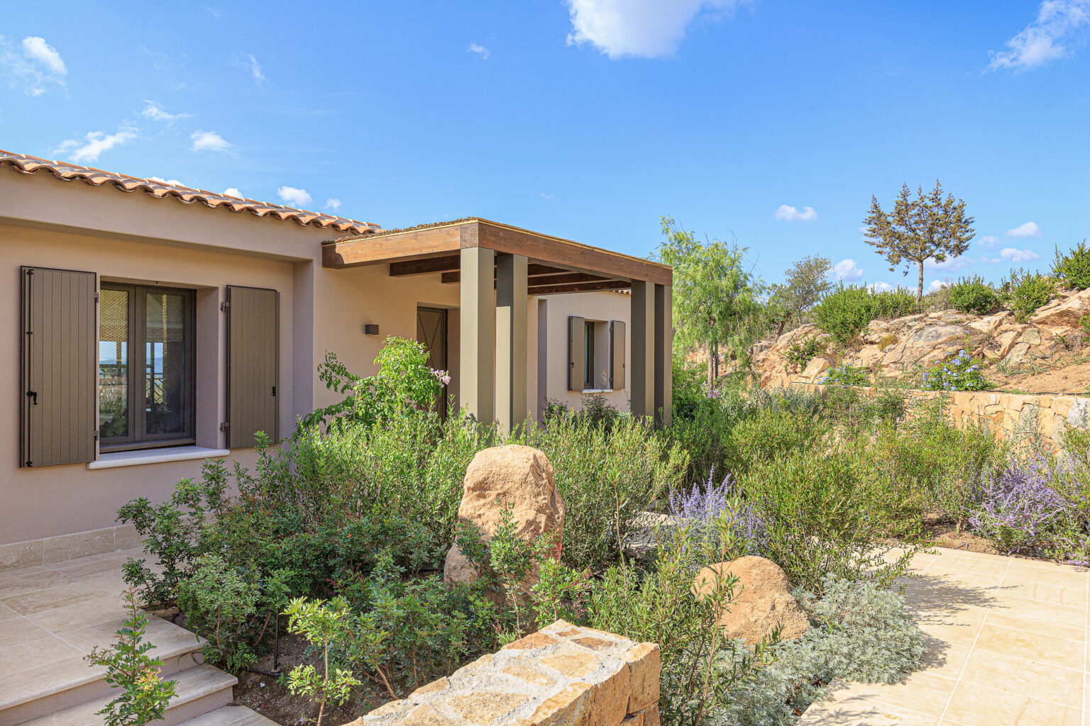 Charming modern house exterior with a covered porch, surrounded by lush greenery and flowering plants under a bright blue sky. Ideal for showcasing outdoor living spaces and landscaping design. | Only on SopranoVillas