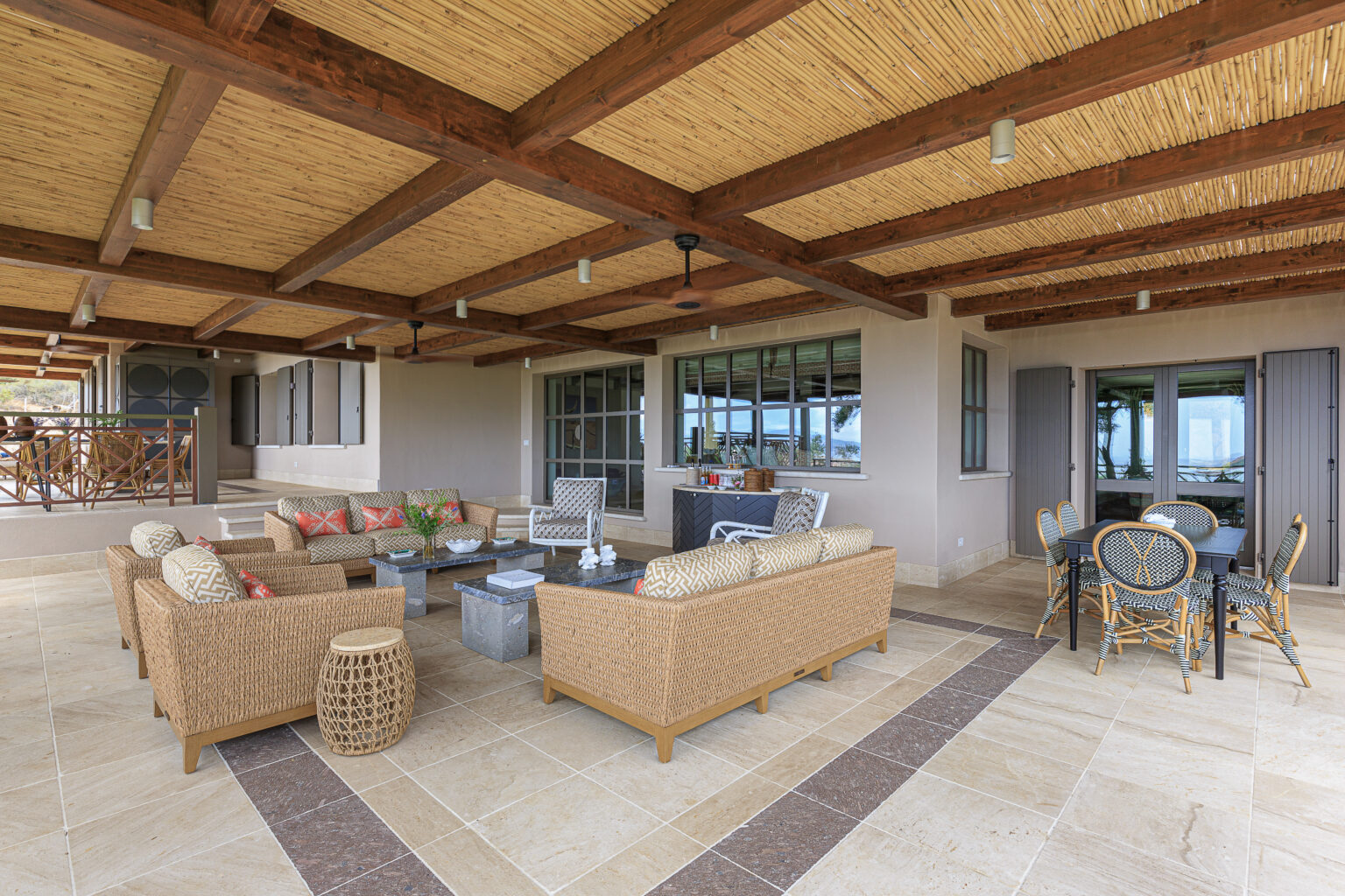 Spacious outdoor living area featuring comfortable wicker furniture with colorful cushions, a stone coffee table, and a dining set, all under a wooden beamed ceiling, ideal for relaxation and entertainment. | Only on SopranoVillas