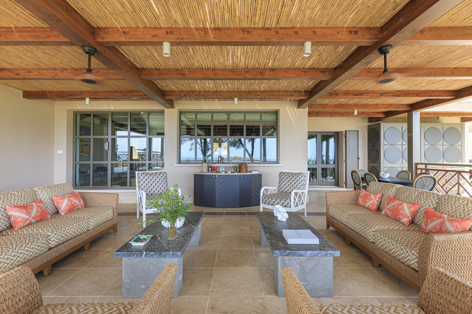 Spacious outdoor living area featuring comfortable wicker seating with decorative pillows, stone coffee tables, and a stylish bar cabinet, all under a bamboo ceiling. Large windows provide scenic views of the surrounding landscape, creating a perfect space for relaxation and entertainment. | Only on SopranoVillas