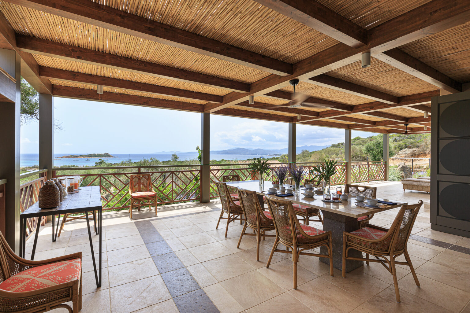 Spacious outdoor dining area with a stunning ocean view, featuring a long table set for meals, surrounded by stylish wicker chairs and tropical plants, under a wooden pergola. | Only on SopranoVillas