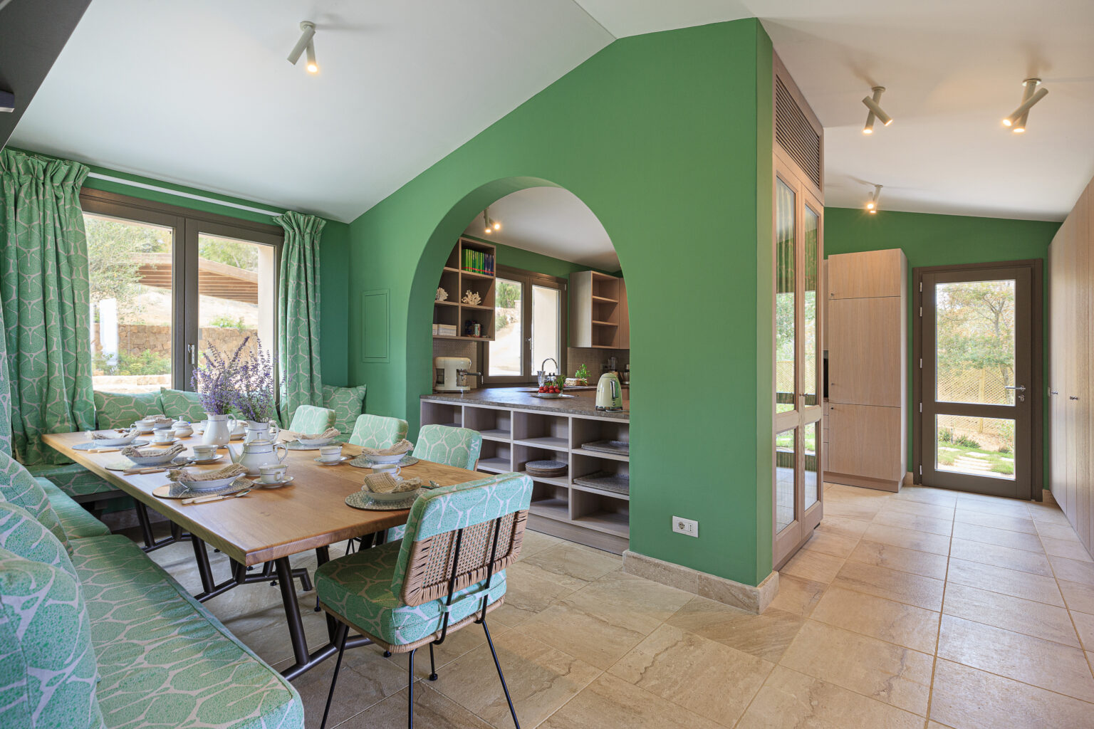 Modern kitchen and dining area featuring vibrant green walls, stylish patterned seating, and a wooden dining table set for a meal. Natural light floods the space through large windows, creating a welcoming atmosphere. The kitchen includes open shelving and contemporary appliances, enhancing the overall aesthetic. | Only on SopranoVillas