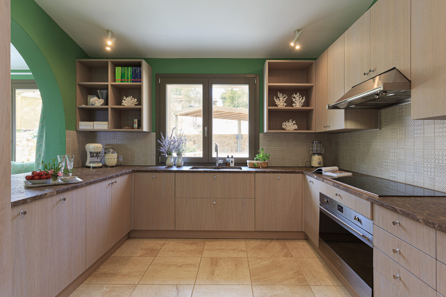 Modern kitchen design featuring light wood cabinetry, green accent walls, and a spacious countertop. The kitchen is equipped with a stainless steel oven, a blender, and decorative elements like coral displays and fresh herbs. Natural light streams through a large window, enhancing the inviting atmosphere. | Only on SopranoVillas