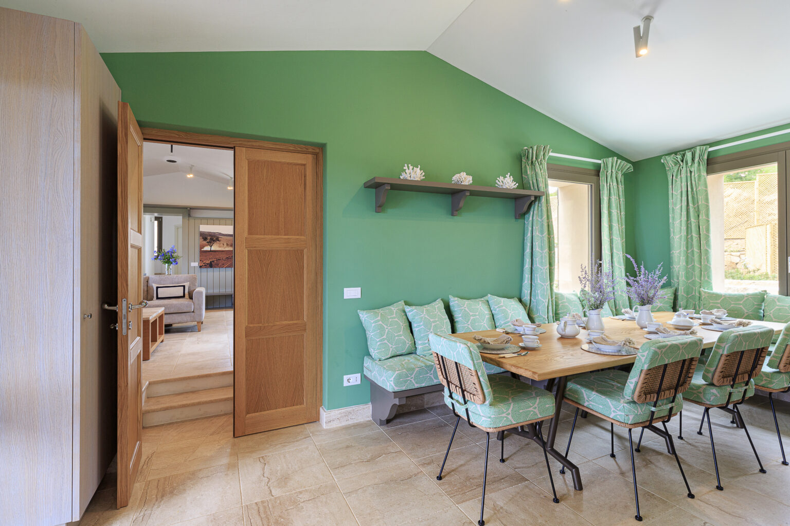 Bright and inviting dining area featuring a green accent wall, a wooden table set for a meal, and cushioned seating. The room is illuminated by natural light from large windows, complemented by decorative curtains and a modern door leading to another space. | Only on SopranoVillas