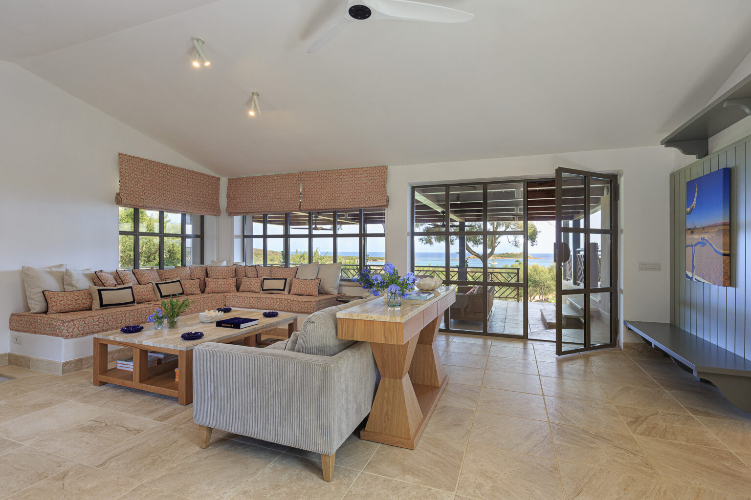 Spacious and stylish living room featuring a cozy sectional sofa with decorative cushions, a wooden coffee table, and large windows that offer a scenic view of the outdoors. The room is adorned with modern decor, including a vase of blue flowers, and has access to a patio area. Natural light floods the space, highlighting the elegant tile flooring and ceiling fan. | Only on SopranoVillas