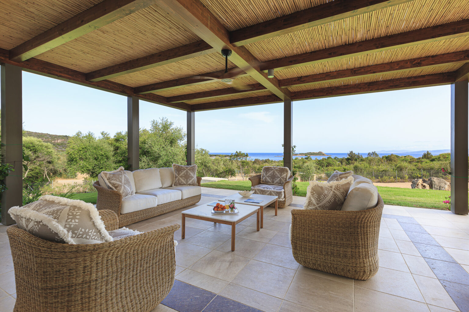 Cozy outdoor living space with a thatched roof, featuring a comfortable wicker sofa and armchairs adorned with decorative pillows, a wooden coffee table with fresh fruits, and a scenic view of lush greenery and the ocean in the background. Ideal for relaxation and entertaining. | Only on SopranoVillas
