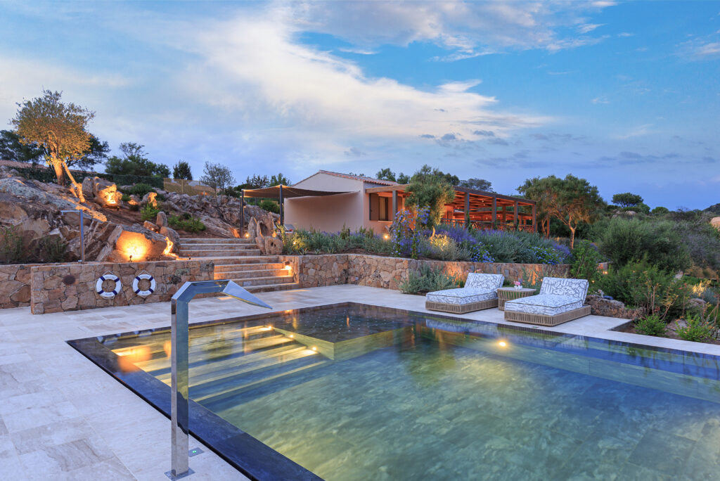 Stunning outdoor pool area featuring modern lounge chairs, surrounded by lush greenery and natural rock formations, with a beautifully designed home in the background under a twilight sky. Ideal for luxury relaxation and outdoor living. | Only on SopranoVillas