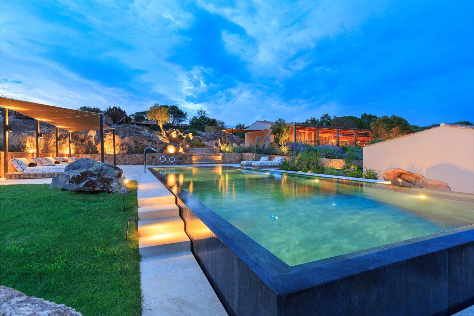 Luxurious outdoor pool surrounded by lush greenery and rock formations, featuring comfortable lounge seating and ambient lighting under a twilight sky. Ideal for relaxation and entertaining. | Only on SopranoVillas